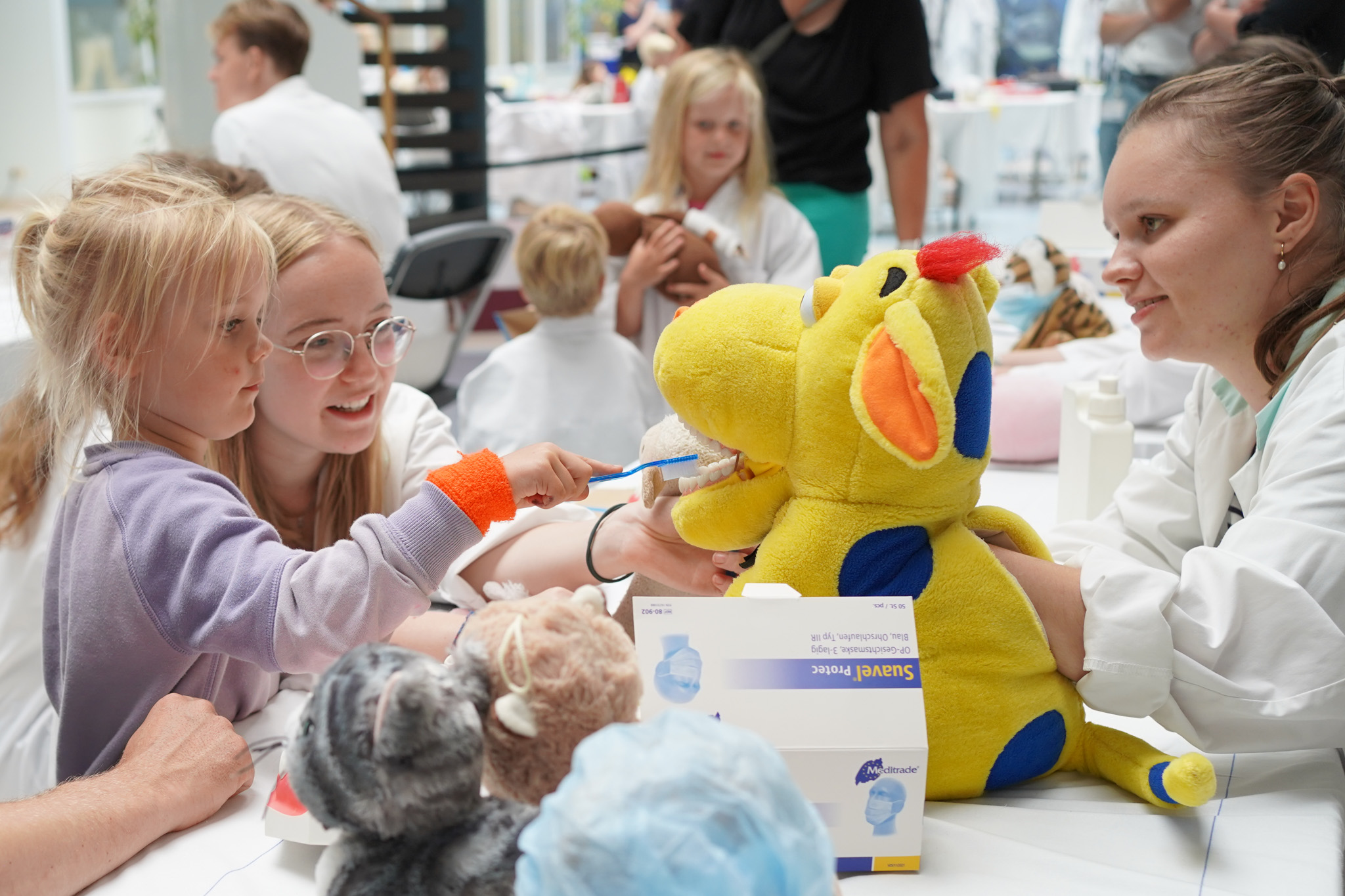 De tanden van een gele knuffelnijlpaard worden gepoetst door een meisje. 2 dokters zitten daarbij