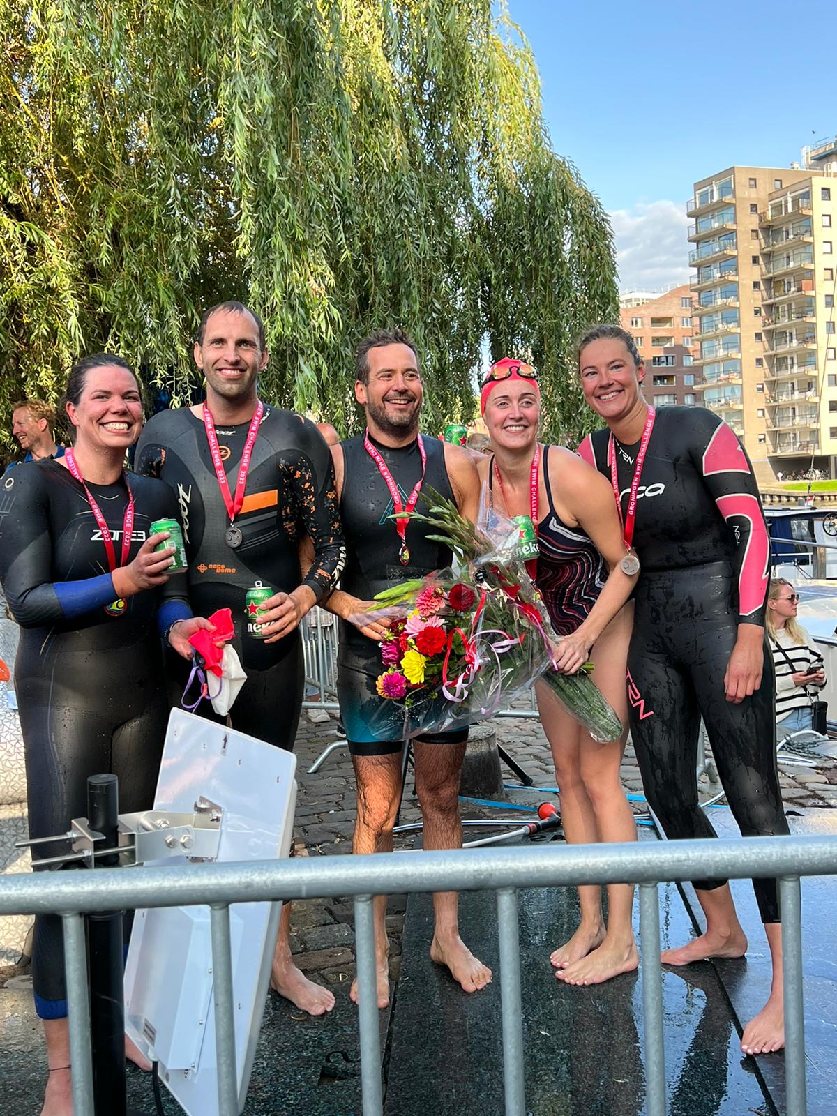 Blij zwemteam 'Van de Baan'. Nog nat van het water juichen ze met bossen bloemen, biertjes en medailles in de hand. Ze hebben hun wetsuit nog aan en staan op de kade.