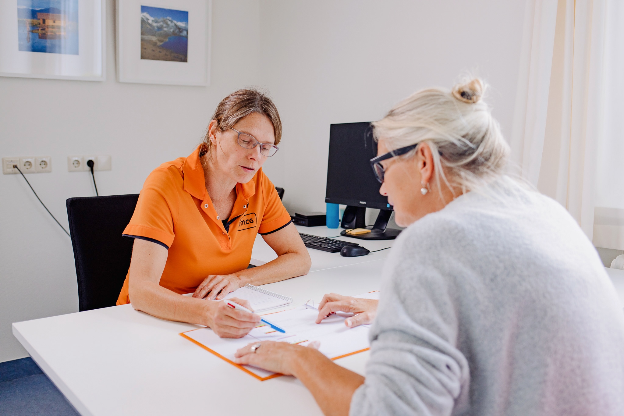 De medewerker van het Centrum voor Revalidatie zit aan een bureau. Ze draagt een oranje polo-shirt met korte mouwen. Tegenoverhaar zit een vrouw met opgestoken, grijs haar. Ze buigt voorover om te kijken naar een plaatje in een boekje. De medewerker wijst het plaatje aan met een pen.